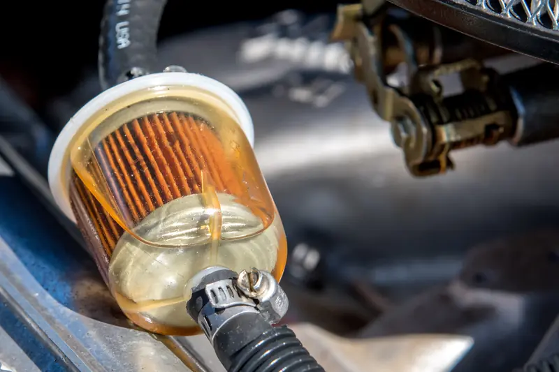 Transparent cylindrical fuel filter with orange-brown pleated filter element visible, connected to black hoses with metal clamps in engine bay