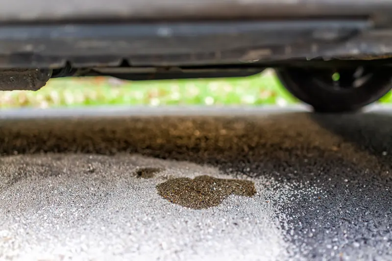 Dark fluid leak puddle on asphalt beneath vehicle undercarriage indicating oil or transmission fluid leak