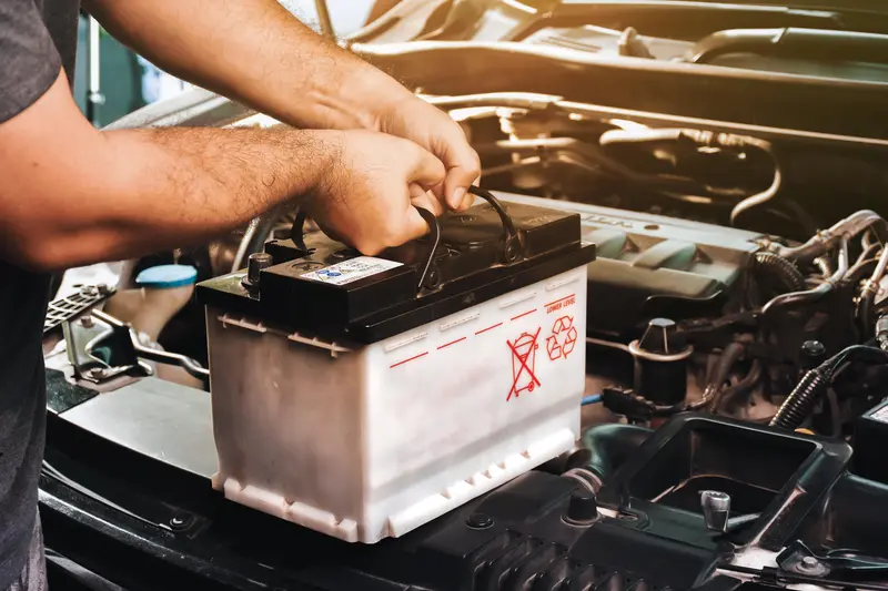 Hands handling white car battery with black top and carrying handle, showing warning symbols, being installed or removed from engine bay with visible hoses and wires