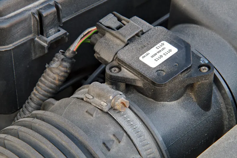 MAF sensor in engine bay showing black plastic housing with white label, electrical connector with wiring harness, and ribbed air intake hose