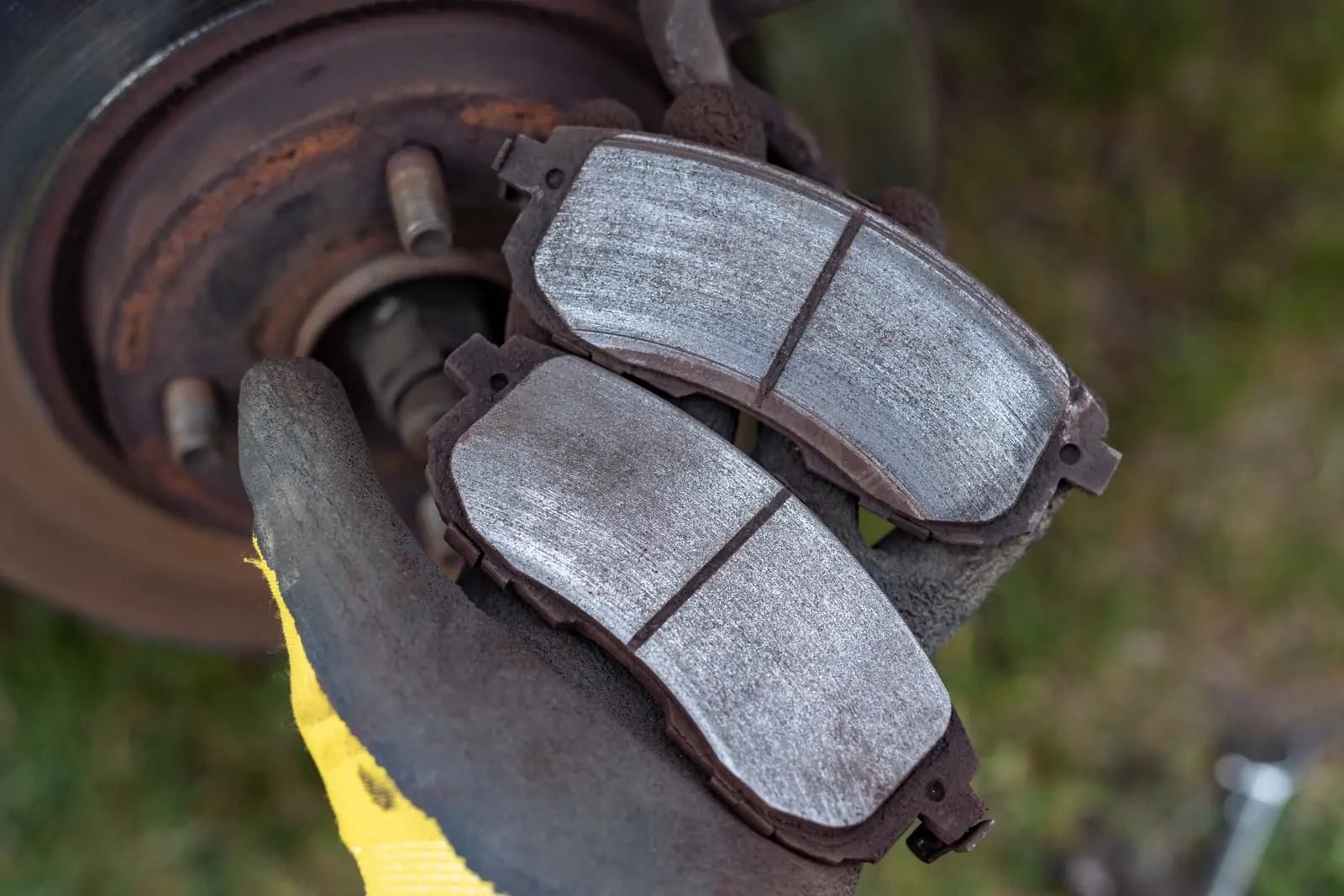 Two new brake pads with light grey friction material and dark backing plates held by gloved hand, with rusty brake rotor visible in background