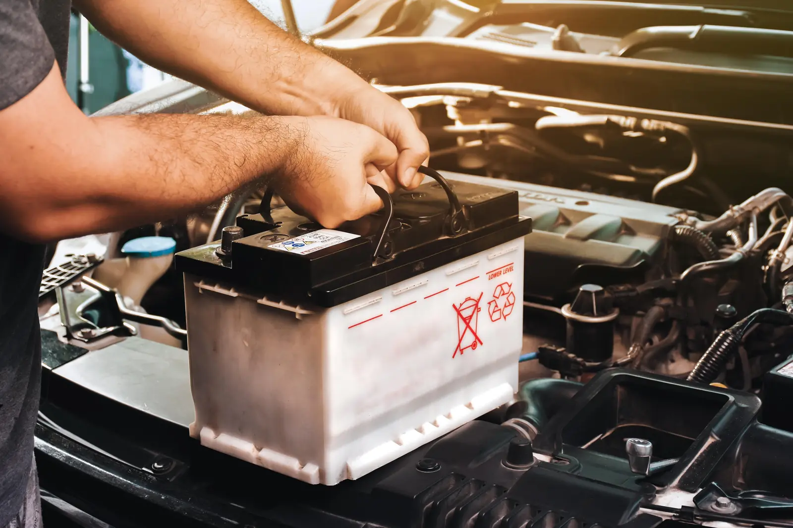 Hands handling white car battery with black top and carrying handle, showing warning symbols, being installed or removed from engine bay with visible hoses and wires