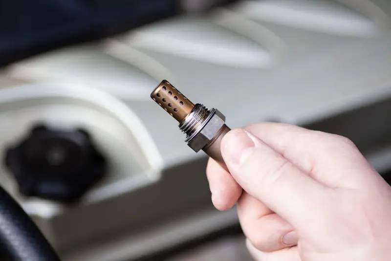 Hand holding oxygen sensor (lambda sensor) showing metallic body with brass-colored perforated tip, hexagonal nut section, and threaded end against engine bay background